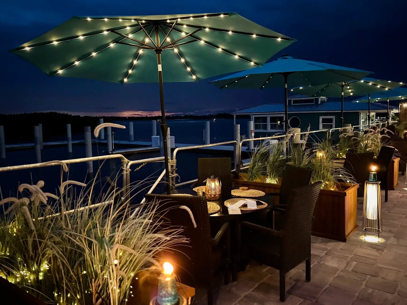 Outdoor waterfront patio with string lights, umbrellas, and sunset views