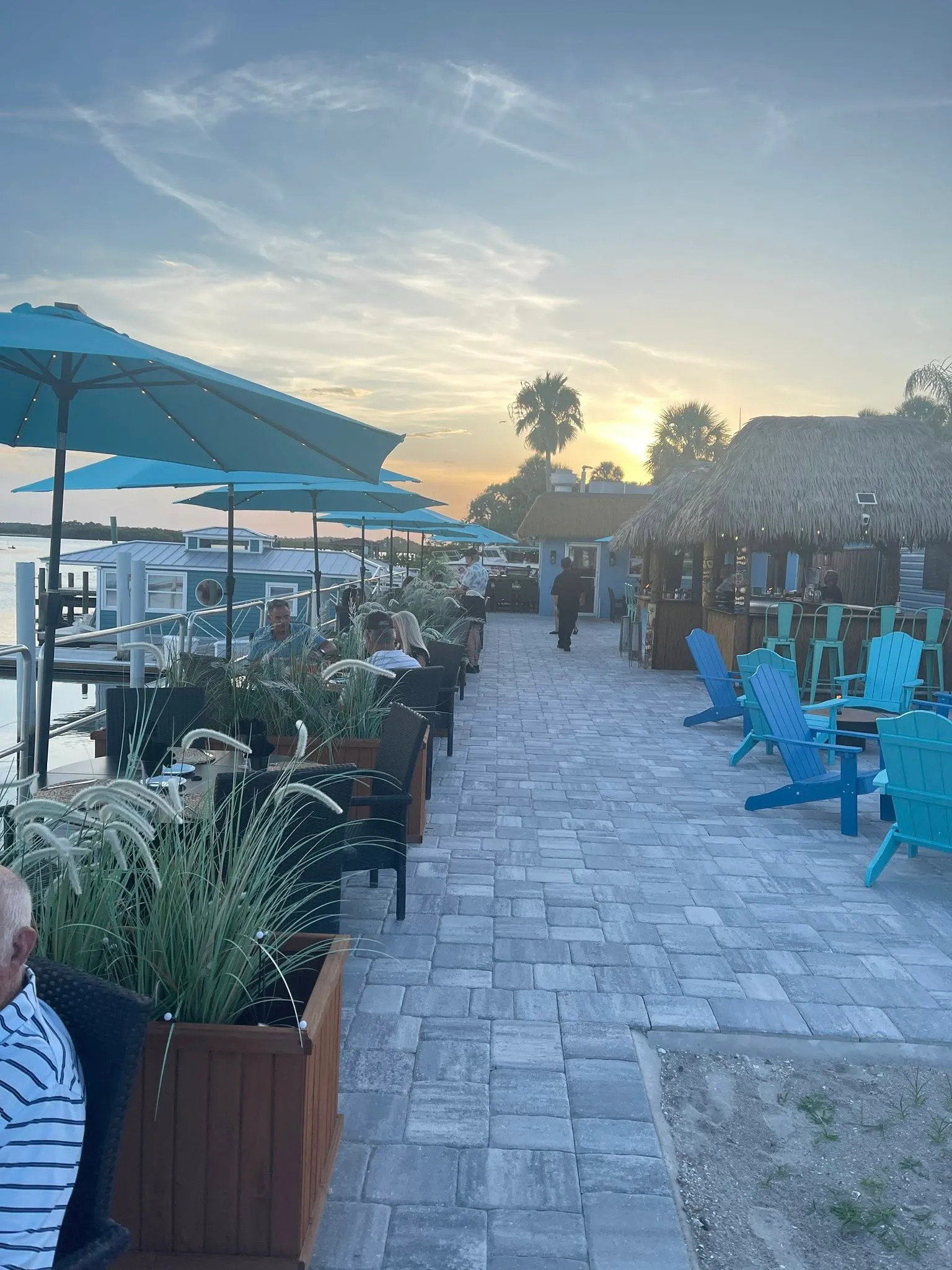Outdoor waterfront dining area at HT's Sand Bar & Bistro with guests seated under blue umbrellas and a tiki-style bar